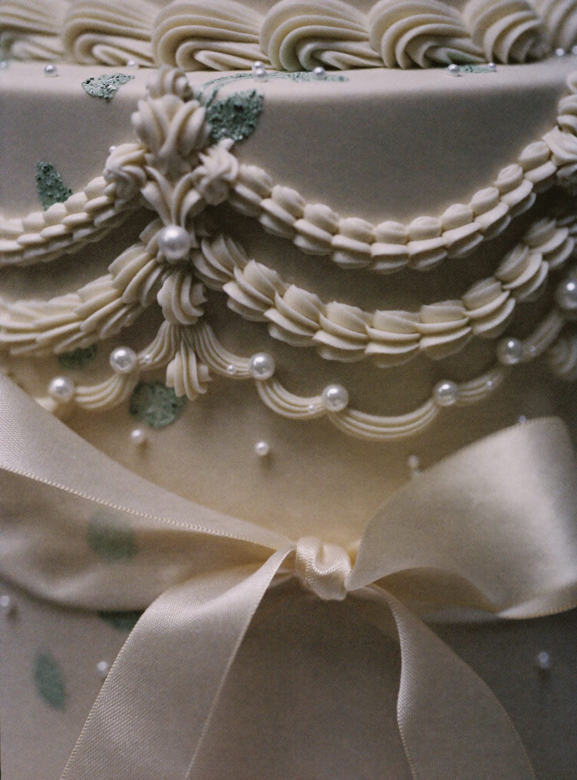 Close up of ivory Lambeth over piping, sugar pearls and painted sage leaves with a butter silk ribbon